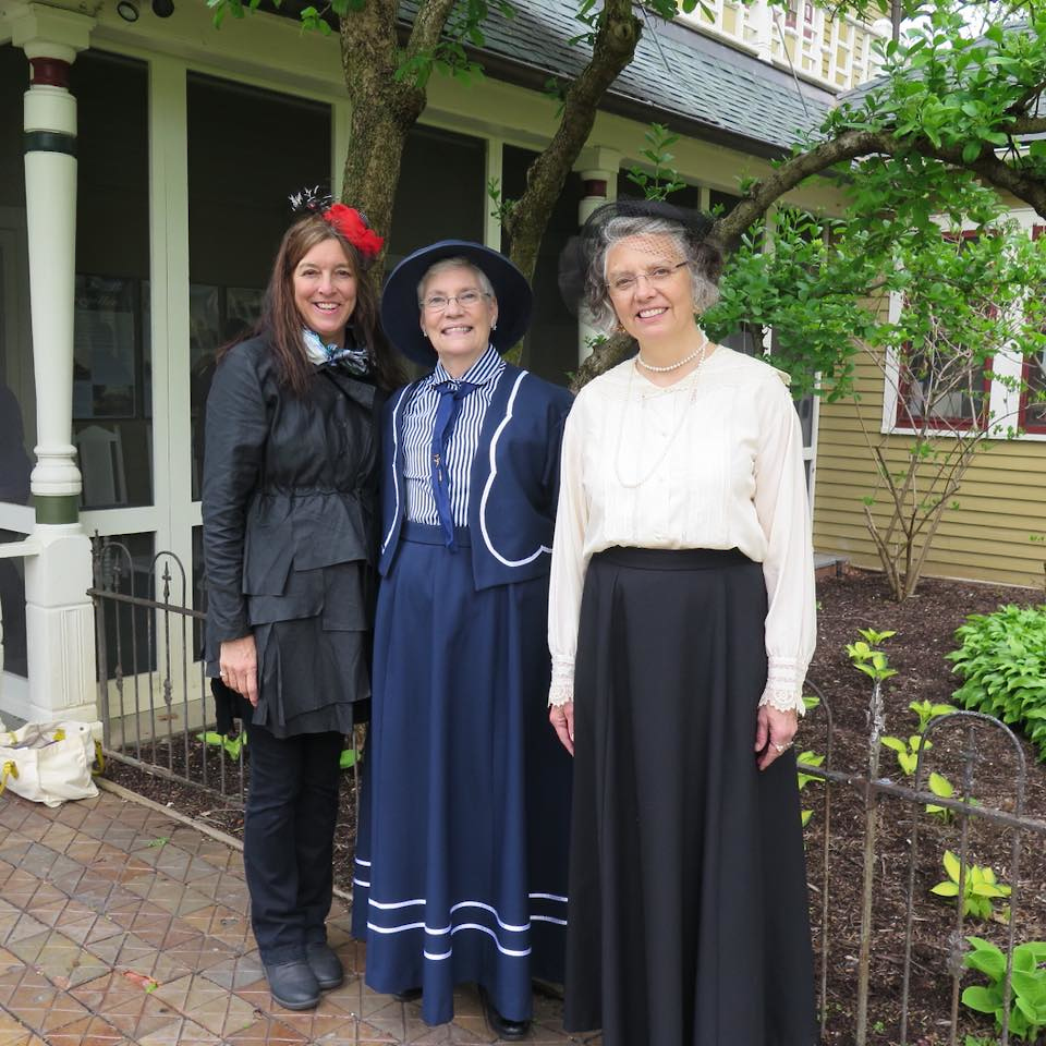 Dressed in period clothes, three white woman smile and pose for the camera during their employment.